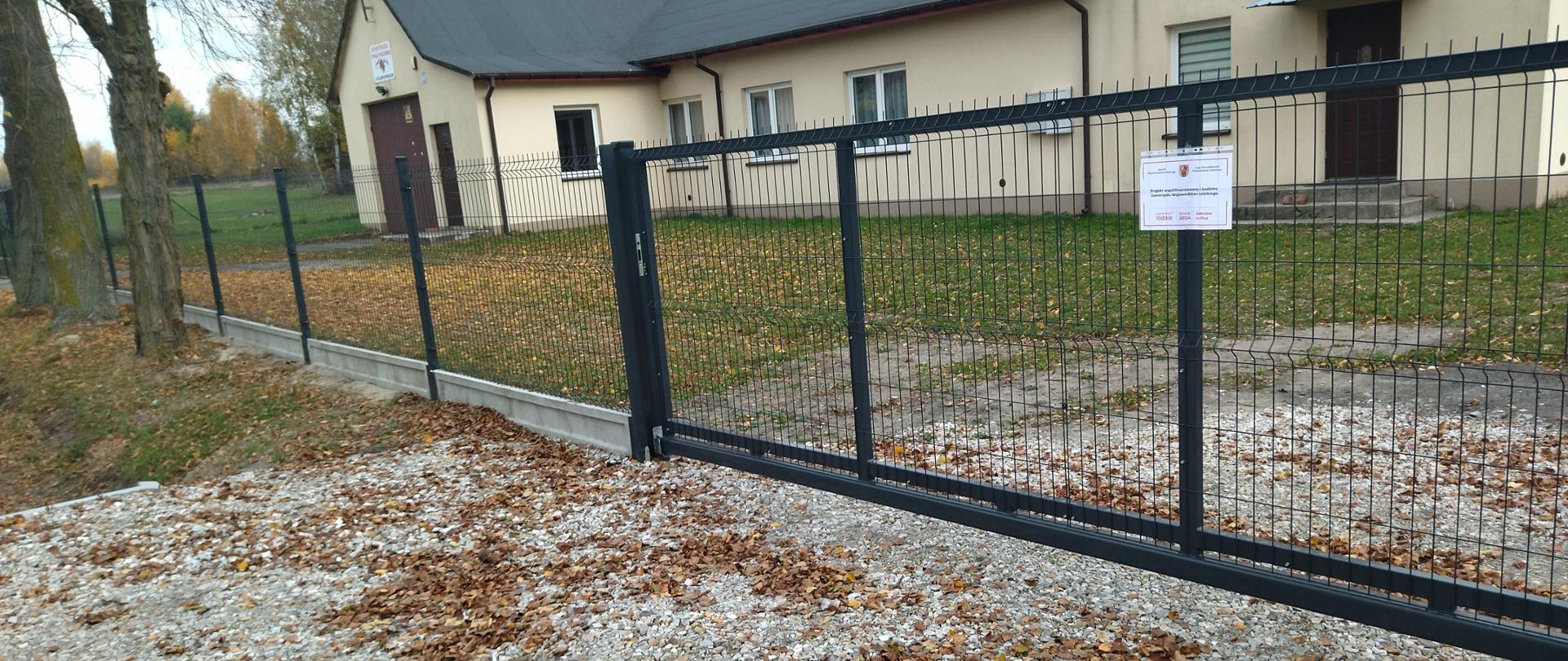 Obraz przedstawia metalowe ogrodzenie z przesuwaną bramą, otaczające teren świetlicy wiejskiej w Ślądkowicach. Na ogrodzeniu znajduje się tablica informacyjna z tekstem dotyczącym współfinansowania projektu z budżetu Samorządu Województwa Łódzkiego. Za ogrodzeniem widoczny jest trawiasty teren, kilka drzew oraz budynki w tle. Na ziemi znajdują się opadłe liście, co sugeruje jesienną porę roku.