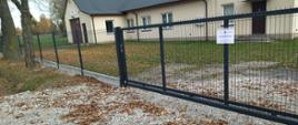 Obraz przedstawia metalowe ogrodzenie z przesuwaną bramą, otaczające teren świetlicy wiejskiej w Ślądkowicach. Na ogrodzeniu znajduje się tablica informacyjna z tekstem dotyczącym współfinansowania projektu z budżetu Samorządu Województwa Łódzkiego. Za ogrodzeniem widoczny jest trawiasty teren, kilka drzew oraz budynki w tle. Na ziemi znajdują się opadłe liście, co sugeruje jesienną porę roku.