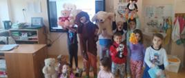 Przedszkolaki w swojej sali stoją z uniesionymi do góry misiami, w tle tablica interaktywna z prezentacją związaną z misiem