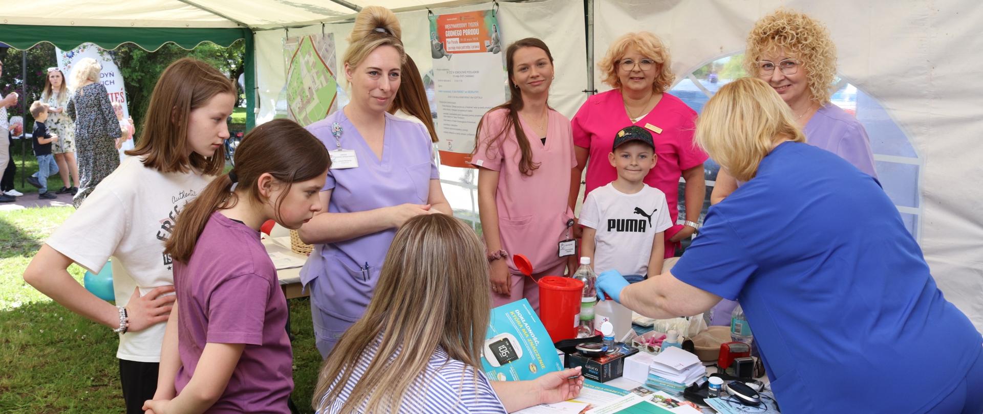 Pod białym namiotem widzimy stanowisko, przy którym pielęgniarki wykonują badania profilaktyczne.