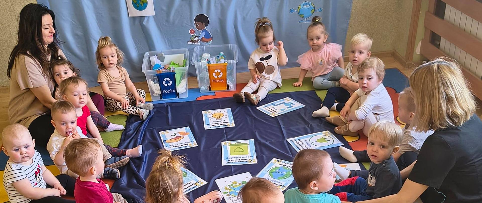 Na zdjęciu są dzieci i panie z grupy Pszczółki. Wszyscy siedzą w kole. Na środku leży granatowy materiał a na nim ilustracje tematyczne. W tle , na błękitnym materiale widnieje napis : "Dzień Ziemi"