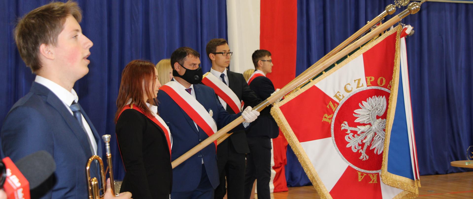 Ludzie stoją w pozycji na baczność. Mężczyzna trzyma trąbkę, a inny opuszczony sztandar. 