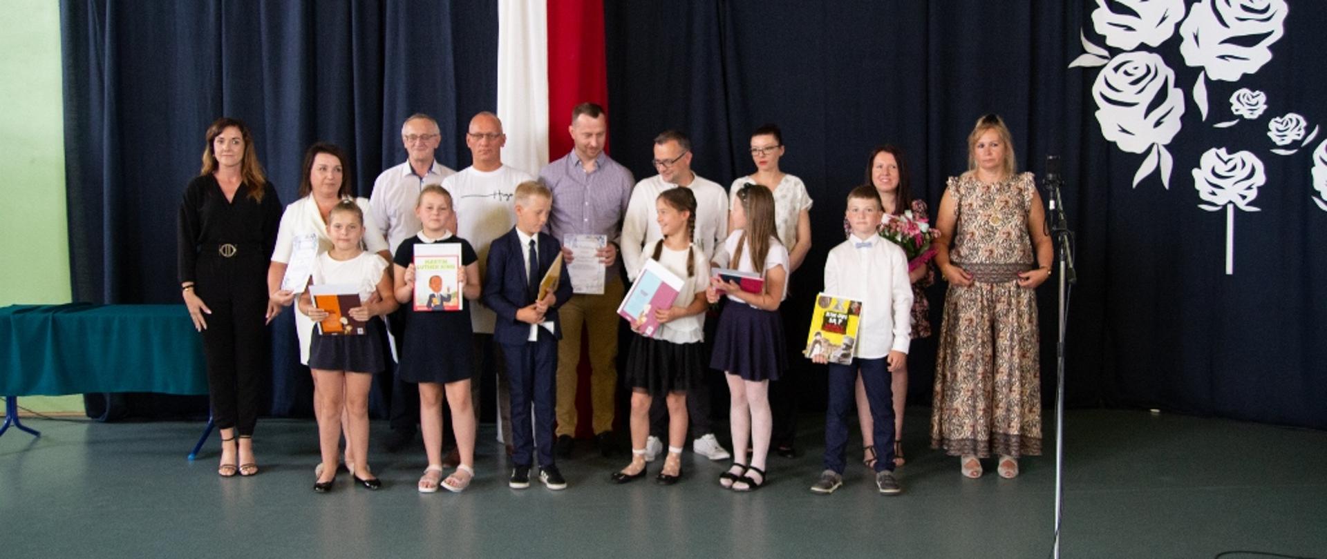 Dorośli stojący w rzędzie, przed nimi stoją dzieci z dyplomami w rękach