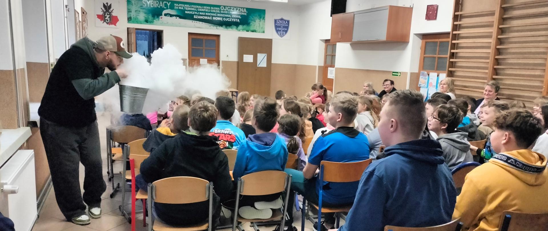 Pan ubrany w czarno-szarą bluzę i czapkę z daszkiem trzyma w dłoniach metalowe wiaderko, z którego wydobywa się dym. Mężczyzna przechadza się wokół uczniów siedzących na krzesełkach.