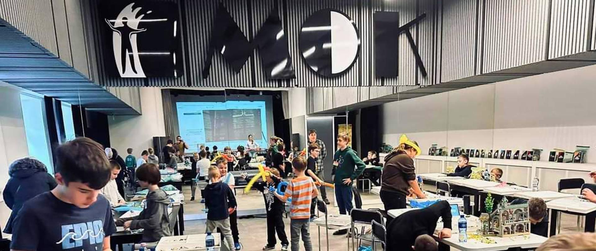 Dzieci i młodzież pracujące przy stołach z klockami LEGO podczas warsztatów w sali Miejskiego Ośrodka Kultury; w tle inni uczestnicy i opiekunowie.