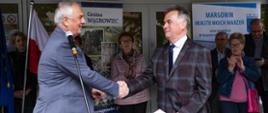 Burmistrz Margonina Janusz Piechocki ściskający dłoń z posłem Grzegorzem Piechowiakiem przed budynkiem przystanku autobusowego w Margoninie, w tle osoby, banery gminy Margonin i Wągrowiec, flagi polski i UE