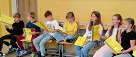 Dzieci siedzą w kole w klasie szkolnej i czytają założenia programu z żółtych kartek