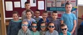 Ubrani na niebiesko uczniowie w niebieskich okularach na nosie stoją lub kucają na szkolnym korytarzu, za nimi gazetka informacyjna na temat praw dziecka ozdobiona wyciętymi z papieru kolorowymi balonami