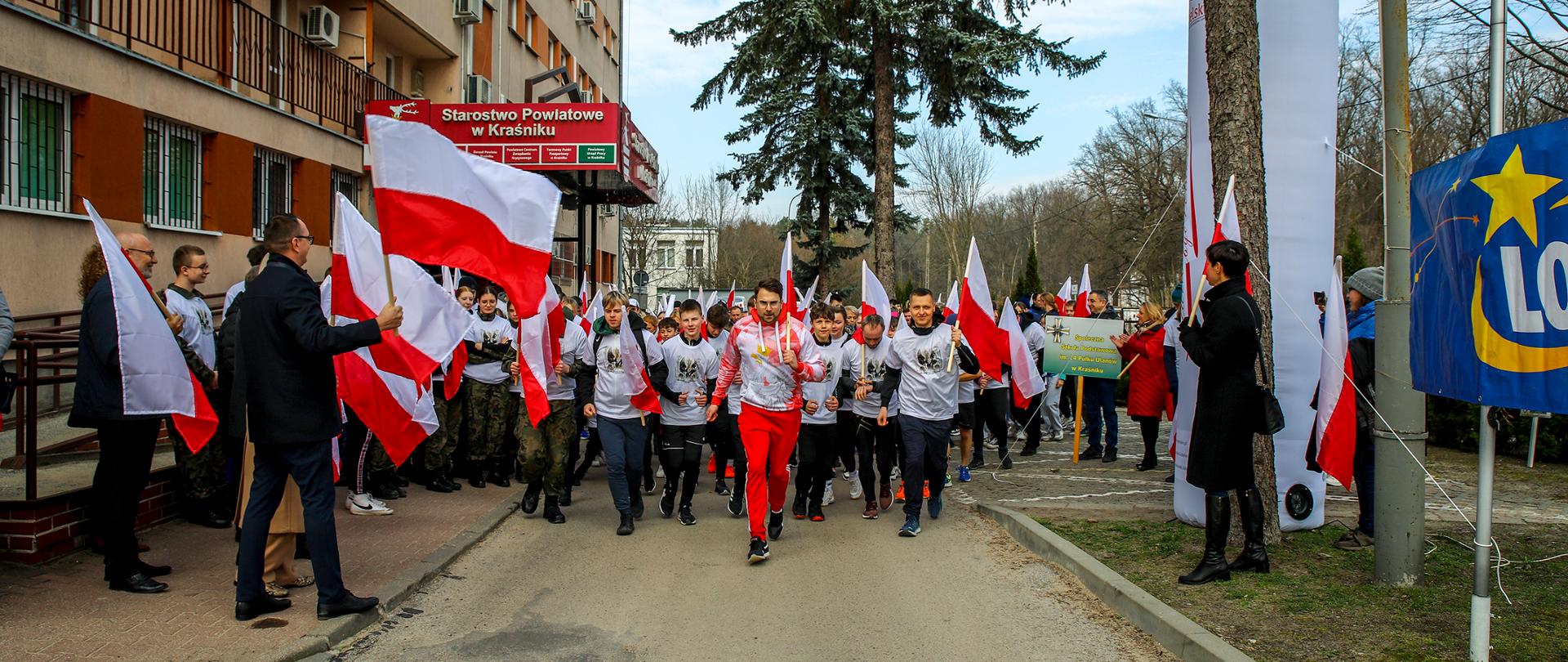 Na zdjęciu grupa ludzi podczas startu biegu. Każdy z uczestników ubrany jest w białą koszulkę z wizerunkiem Majora Hieronima Dekutowskiego oraz trzyma biało-czerwoną flagę.