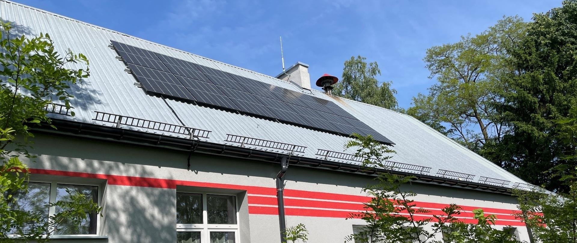 Instalacja fotowoltaiczna na szarym dachu budynku remizy OSP Udrzyn. Wokół drzewa.