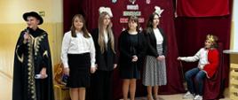 Uczniowie klasy ósmej prezentują program artystyczny na korytarzu szkolnym, jeden uczeń przebrany za króla w czerwonym płaszczu i koronie na głowie, drugi uczeń w czarnej szacie poety-wieszcza, w tle na bordowej zasłonie dekoracja związana z okazją: biało-czerwona flaga oraz wycięte z papieru: otwarta książka, czapka absolwenta i pióro, przed uczniami pani dyrektor szkoły przemawia do zgromadzonych dzieci