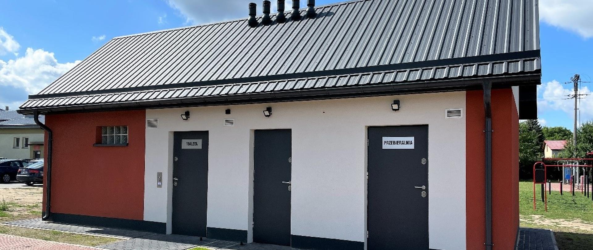 Budynek z biało-pomarańczową elewacją i grafitowym dachem z blachy trapezowej. w budynku są 3 pary drzwi i okno.