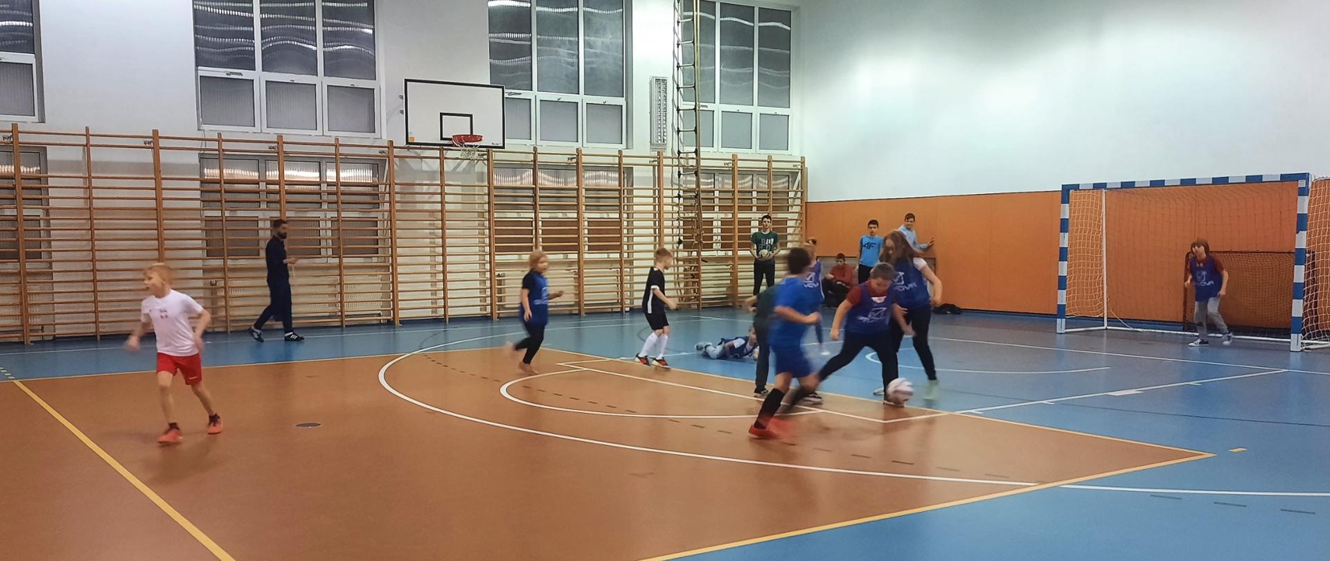 Zdjęcie przedstawia grupkę dzieci grających w piłkę nożną na hali sportowej.