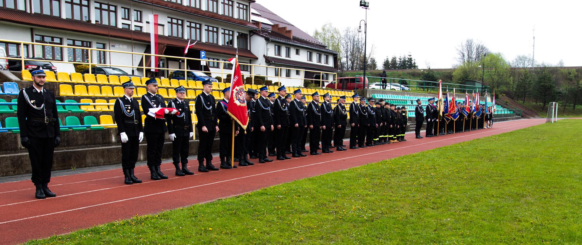 Na boisku sportowym w rzędzie stoją druhowie z OSP z pocztami sztandarowymi