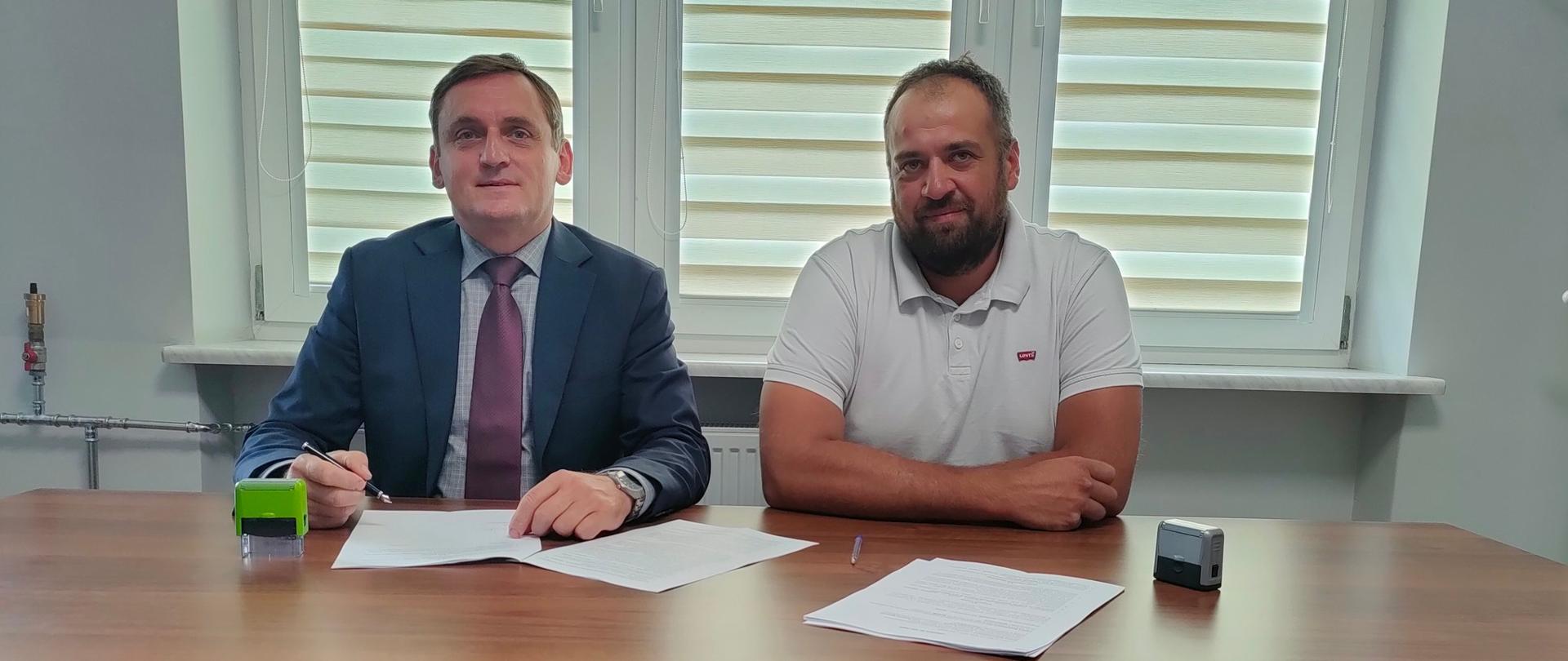 Zdjęcie przedstawia Dwóch mężczyzn siedzi przy stole i podpisuje dokumenty.