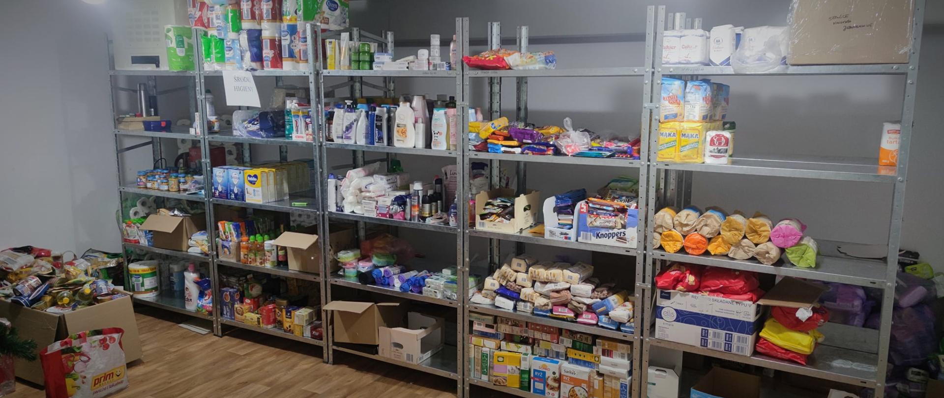 Pomoc humanitarna zorganizowana przez Powiat Przasnyski dla obywateli Ukrainy. Na zdjęciu magazyn z półkami wypełnionymi artykułami spożywczymi i chemii gospodarczej. Na podłodze po lewej stronie zapakowane torby i kartony gotowe do przekazania potrzebującym. 