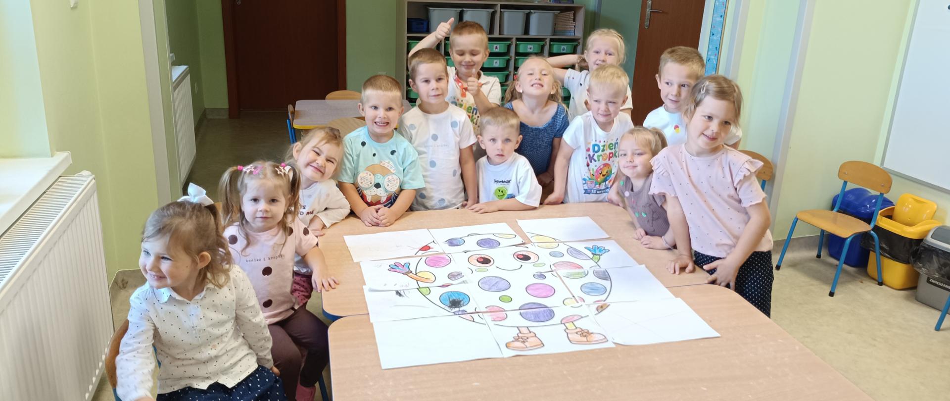 Grupa dzieci przedszkolnych w sali przedszkolnej, po lewej okno, za dziećmi drzwi i tablica korkowa. Część dzieci stoi, część siedzi na małych krzesłach. Na stoliku przed nimi leży kolorowy plakat przedstawiający dużą kropkę. 