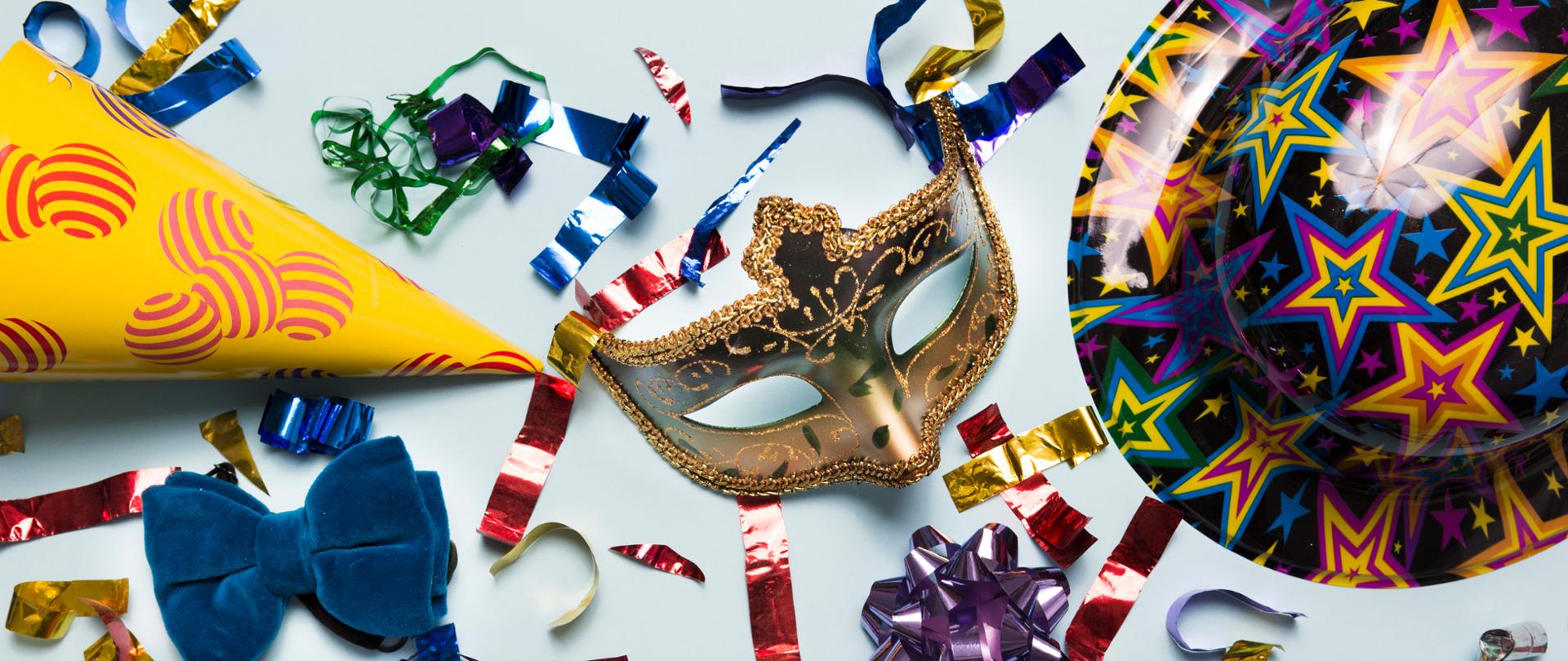 Kolorowe akcesoria karnawałowe rozłożone na jasnym tle: złota maska balowa, papierowa czapka imprezowa, niebieska muszka, balon w gwiazdki oraz kolorowe serpentyny i konfetti, tworzące radosny, świąteczny klimat.