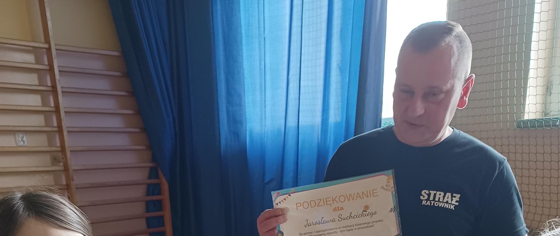 Mężczyzna w koszulce z napisem straż trzyma w rękach kartkę papieru. Przed nim stoją 2 dziewczynki
