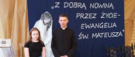 Uczennica klasy siódmej wraz z księdzem uczącym religii pozuje do zdjęcia na tle granatowej dekoracji z białym napisem "Z Dobrą Nowiną przez Życie - Ewangelia Św. Mateusza"