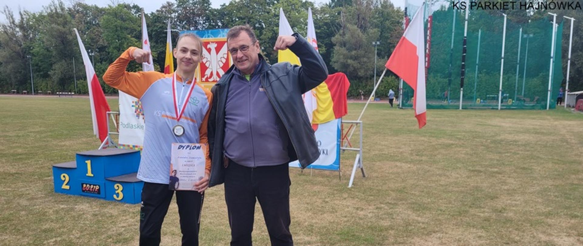 Aleksander z medalem i dyplomem pozuje z trenerem, w tle podium