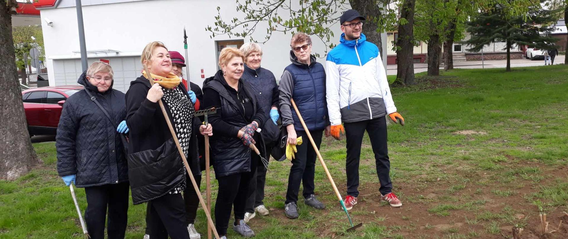 Uczestnicy Klubu Senior+ wraz z Burmistrzem Dąbrowy Białostockiej pozują do zdjęcia w parku miejskim, w rękach trzymają narzędzia ogrodnicze 