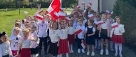 Grupa dzieci spaceruje ulicami Chorzelowa. Dzieci ubrane w barwy narodowe, trzymają w rękach flagi Polski.