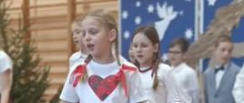 Dziewczynki w białych bluzeczkach i czerwonych góralskich spódnicach w róże prezentują układ taneczny, w tle śpiewają chłopcy w odświętnych strojach