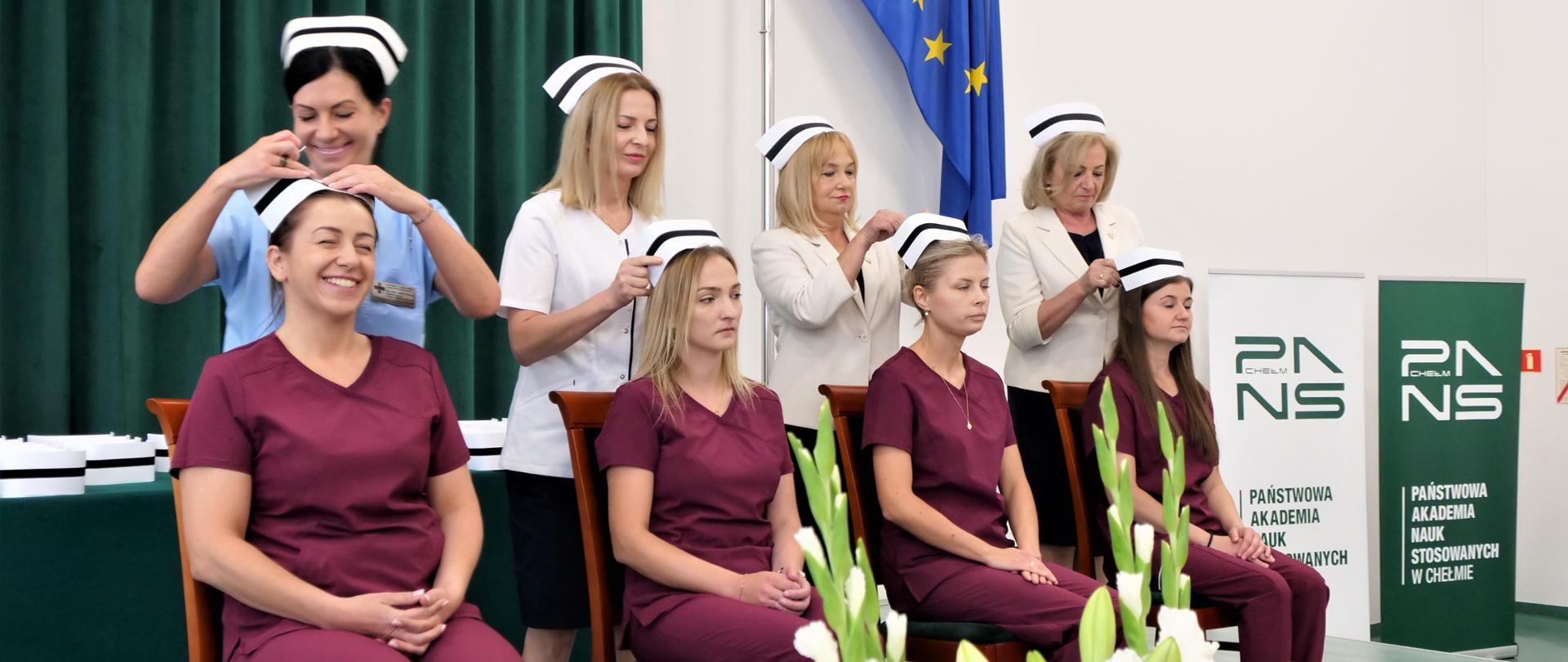 Zdjęcie przedstawia cztery osoby siedzące na krzesłach, a za nimi stoją kolejne cztery, które nakładają im na głowy czepki pielęgniarskie
