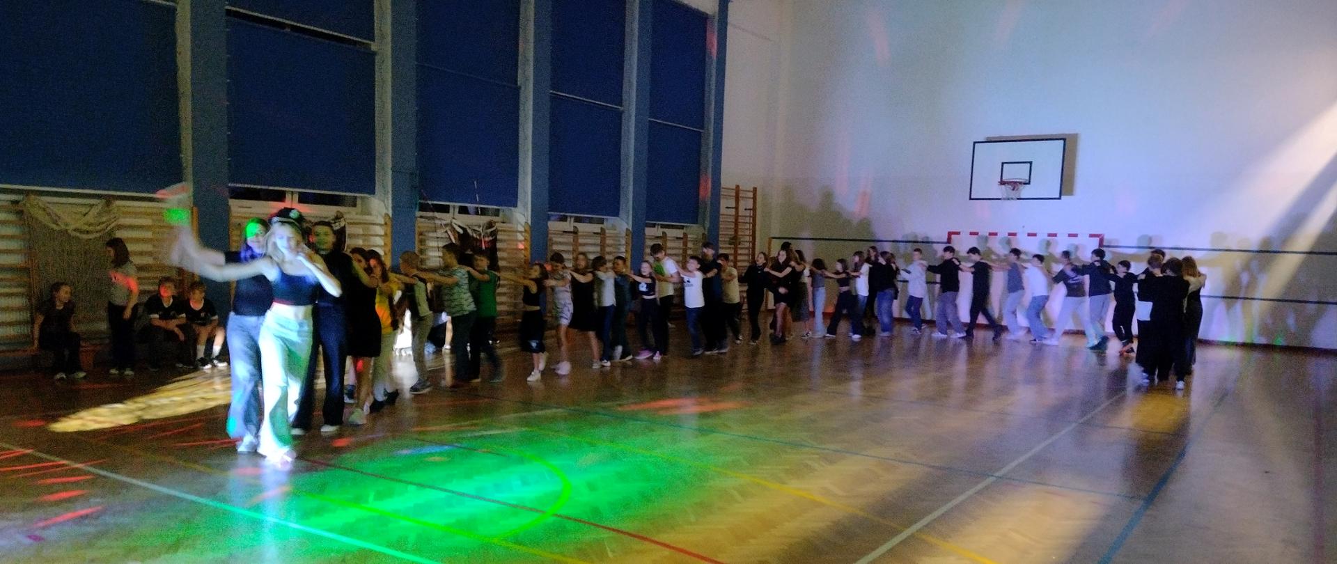 Uczniowie klas IV - VIII tańczą na sali gimnastycznej w rytm muzyki.