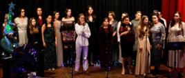 Grupa osób elegancko ubranych na scenie śpiewa wspólnie kolędę, od lewej strony pianista który akompaniuje . Scena udekorowana jest światełkami świątecznymi tło kurtyna czarna.