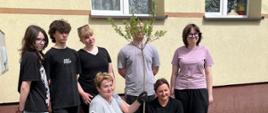 Przed szkołą znajduje się pięcioro uczniów klasy ósmej, ich wychowawczyni oraz Pani Dyrektor. Panie trzymają w dłoniach sadzonkę drzewka. Wszyscy pozują do wspólnego zdjęcia.