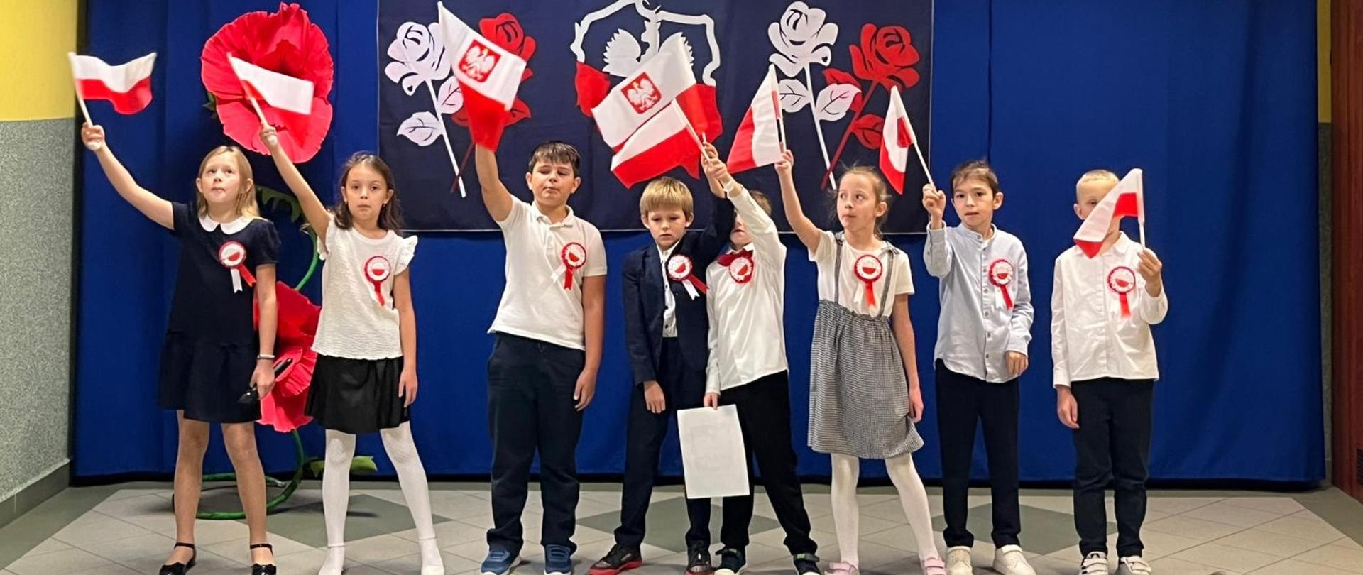 Uczniowie klasy 3 odświętnie ubrani wymachują biało-czerwonymi flagami na akademii, za nimi na niebieskiej kotarze dekoracja z napisem "11 listopada" oraz biało-czerwona mapa Polski z orłem w koronie wewnątrz mapy, po obu stronach biała i czerwona róża