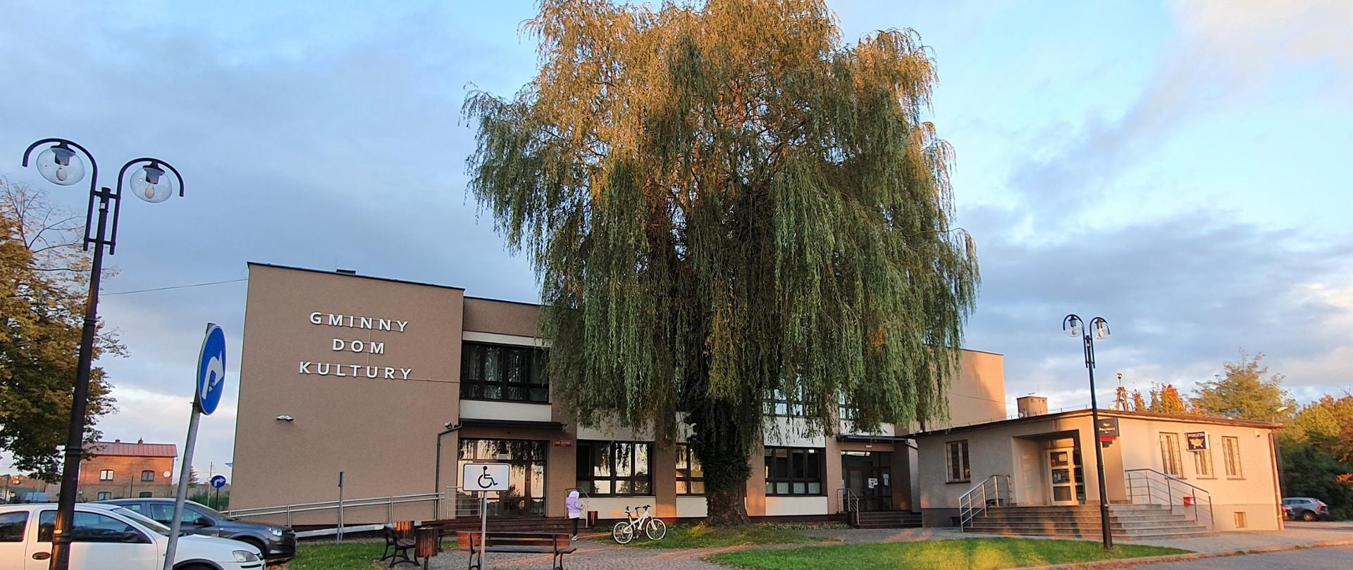 Beżowy budynek z napisem Gminny Dom Kultury a przed nim wysoka zielona wierzba.