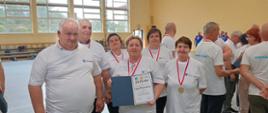 Grupa przedstawicieli Gminy Wietrzychowice w białych koszulkach reprezentacyjnych podczas V Turnieju Sołectw Powiatu Tarnowskiego. Uczestnicy mają na sobie medale na czerwonych wstążkach, a kobieta w centrum trzyma pamiątkowy dyplom w granatowej teczce.