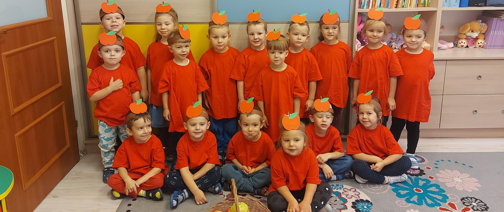 Grupa dzieci w czerwonych koszulkach i z przepaskami na głowach z wzorem jabłka. Przed dziećmi kosz z jabłkami, a nad nimi napis DZIEŃ JABŁKA