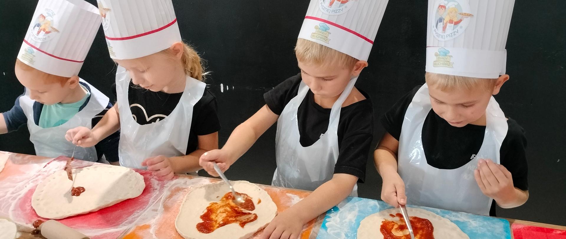 4 dzieci w czapkach kucharskich przygotowuje pizze 