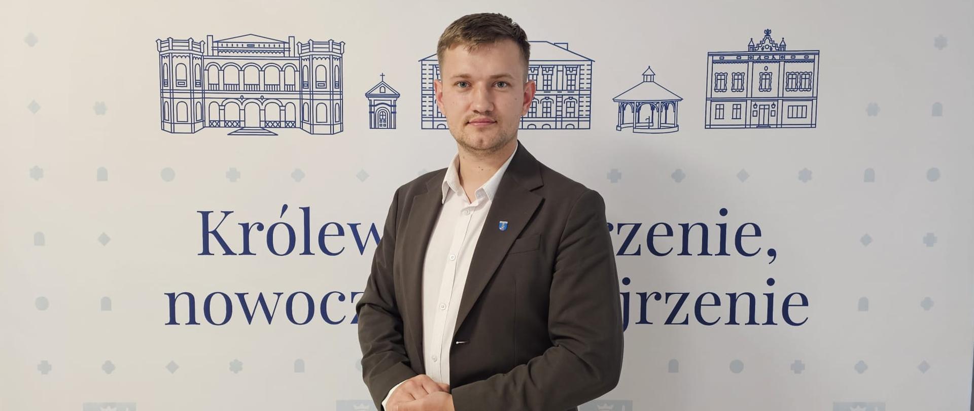 Mężczyzna w ciemnoszarej marynarce stoi na tle ścianki promocyjnej Gminy Tyczyn.
