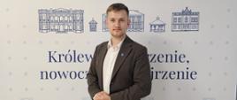 Mężczyzna w ciemnoszarej marynarce stoi na tle ścianki promocyjnej Gminy Tyczyn.
