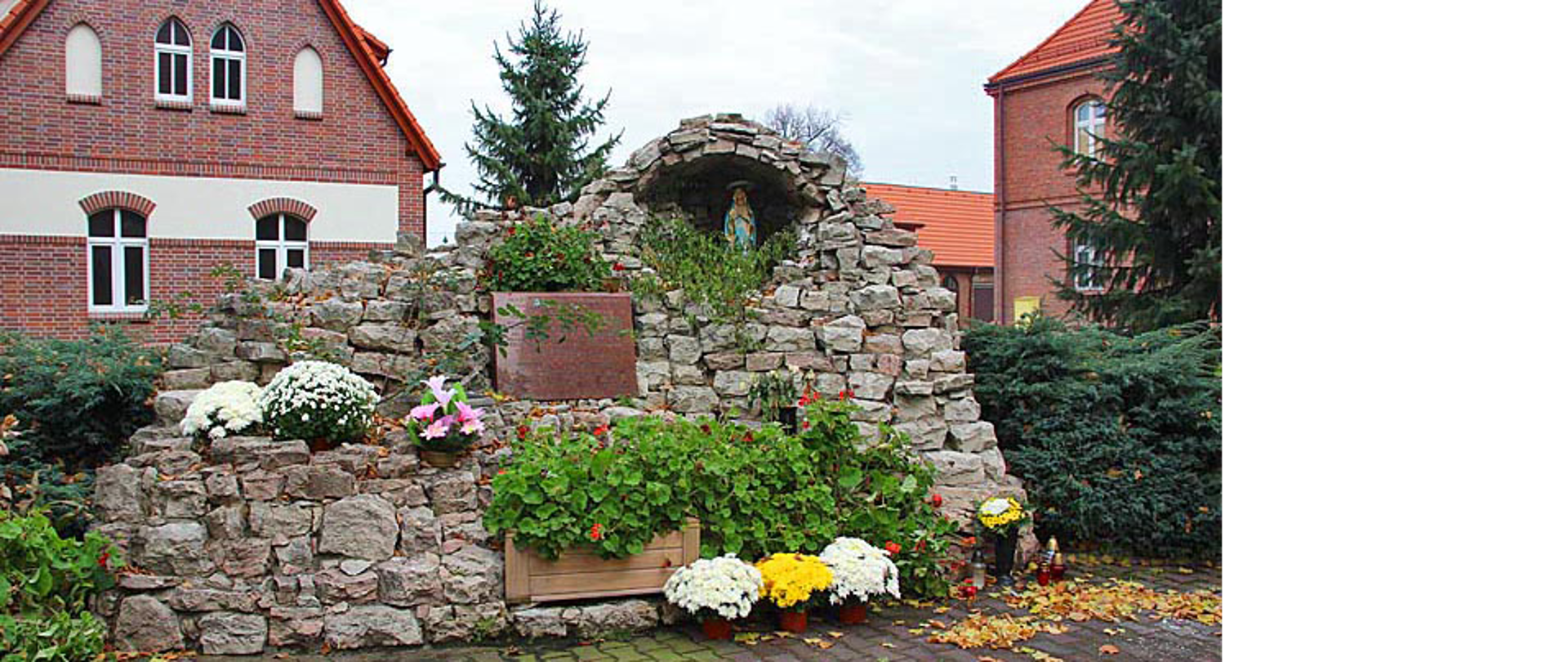 Grota Matki Boskiej z Lourdes utworzona z kamieni, wśród kamieni znajdują się kwiaty oraz tablica informacyjna. W grocie stoi figura Matki Boskiej. W tle widać dom parafialny i budynek probostwa z czerwonej cegły.