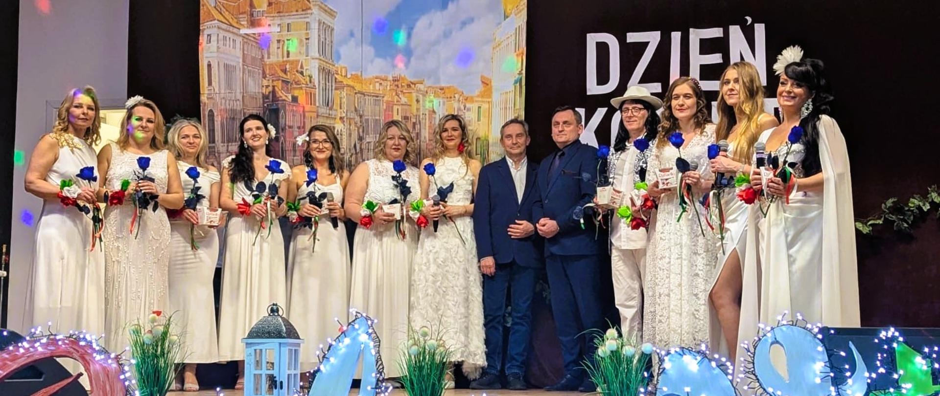 Grupa kobiet ubranych w białe sukienki z niebieskimi różami w dłoniach oraz dwóch mężczyzn ubranych w garnitury pozujących do zdjęcia na scenie