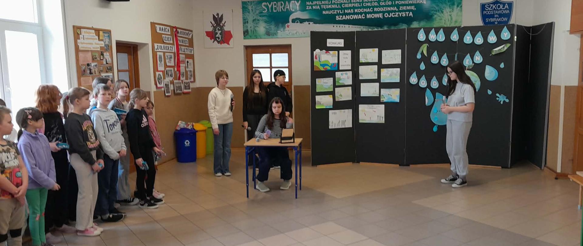 Na scenie znajdują się uczniowie klasy szóstej. Jedna z uczennic siedzi przy stoliku i przygląda się w lusterku. Obok niej stoi czworo uczniów - trzy dziewczyny i chłopak. W dłoniach trzymają mikrofony. Za uczniami, na ściance wiszą plakaty konkursowe na temat wody oraz napis: ŚWIATOWY DZIEŃ WODY.