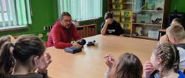 Dzieci siedzą na około dużego kwadratowego stołu. Przemawia do nich mężczyzna w czerwonym swetrze z aparatem fotograficznym trzymanym w rękach