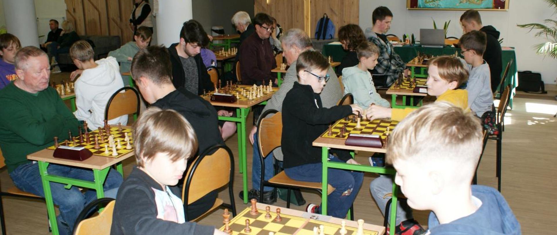 Na zdjęciu widzimy siedzących w sali przy poustawianych obok siebie ławkach zawodników rozgrywających zawody w szachy.