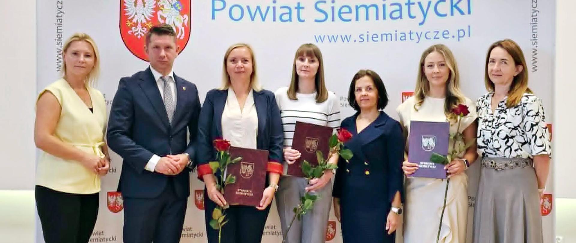 Grupa osób stojących razem przed banerem Powiatu Siemiatyckiego, trzymających certyfikaty i czerwone róże.
