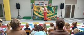 Dzieci z rodzicami siedzą w jasnej sali oglądając występ aktora.