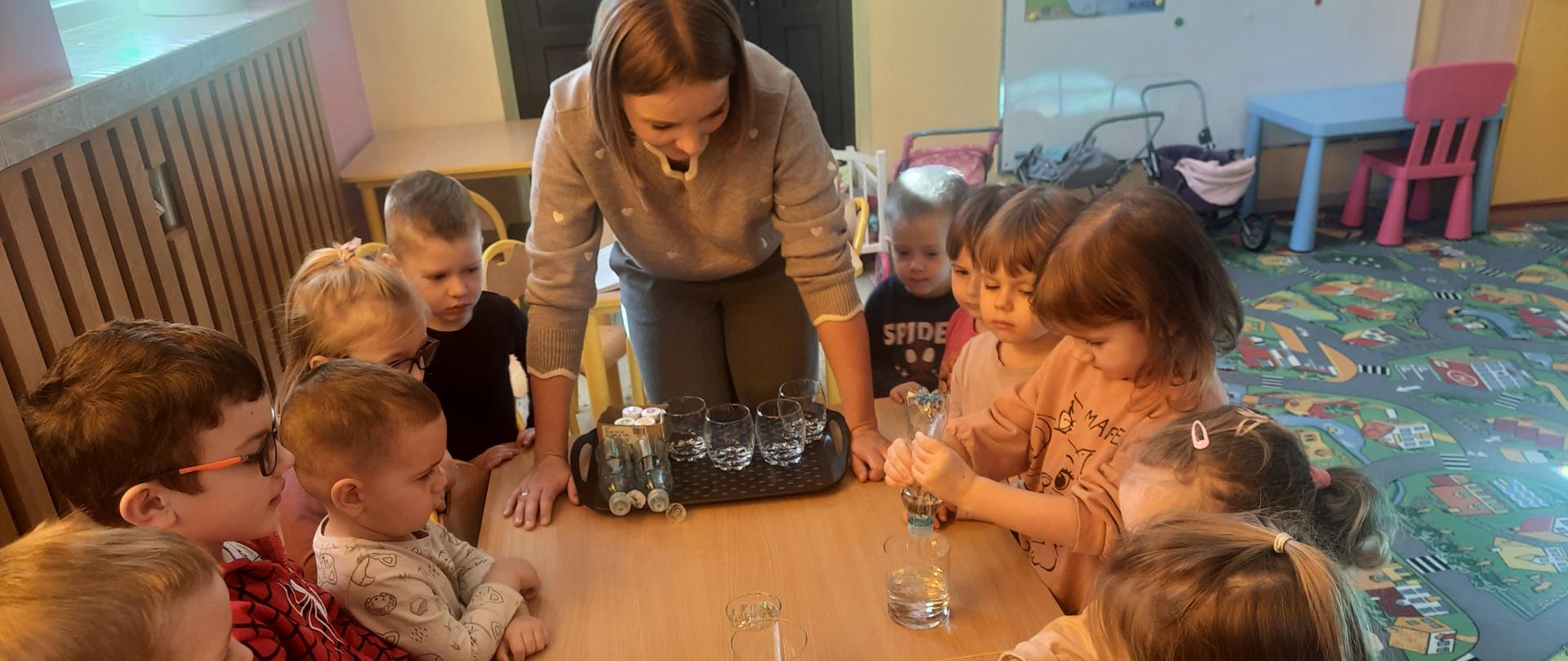Uczniowie zgromadzeni wokół stołu, nauczyciel w centrum, na stole butelki z różnymi płynami.