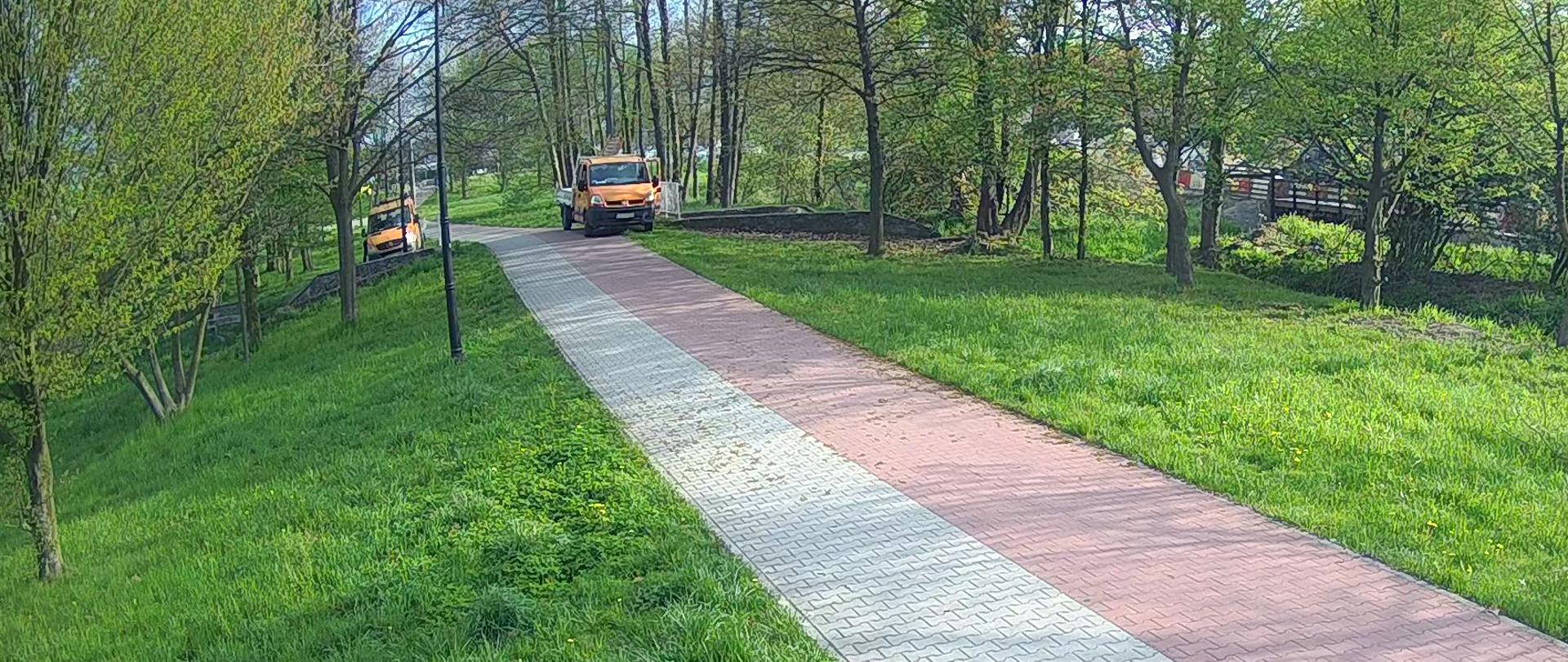 Szaro czerwony chodnik w tle dwa pomarańczowe samochody wśród drzew.