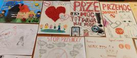 Na zdjęciu widoczny jest stół w sali, na którym ułożono kilkanaście kolorowych prac plastycznych wykonanych przez dzieci. Rysunki przedstawiają hasła i symbole związane z przeciwdziałaniem przemocy, m.in. hasła „STOP przemocy”, „Rodzice nie bijcie dzieci”, „Przemoc to nie moc”. Prace wykorzystują różnorodne techniki – od rysunku kredkami po flamastery – oraz liczne motywy symboliczne, takie jak pęknięte serca, czerwone dłonie, przekreślone znaki czy postacie ilustrujące trudne sytuacje. Całość tworzy kolorową i przejmującą prezentację prac konkursowych o tematyce przeciwdziałania przemocy.