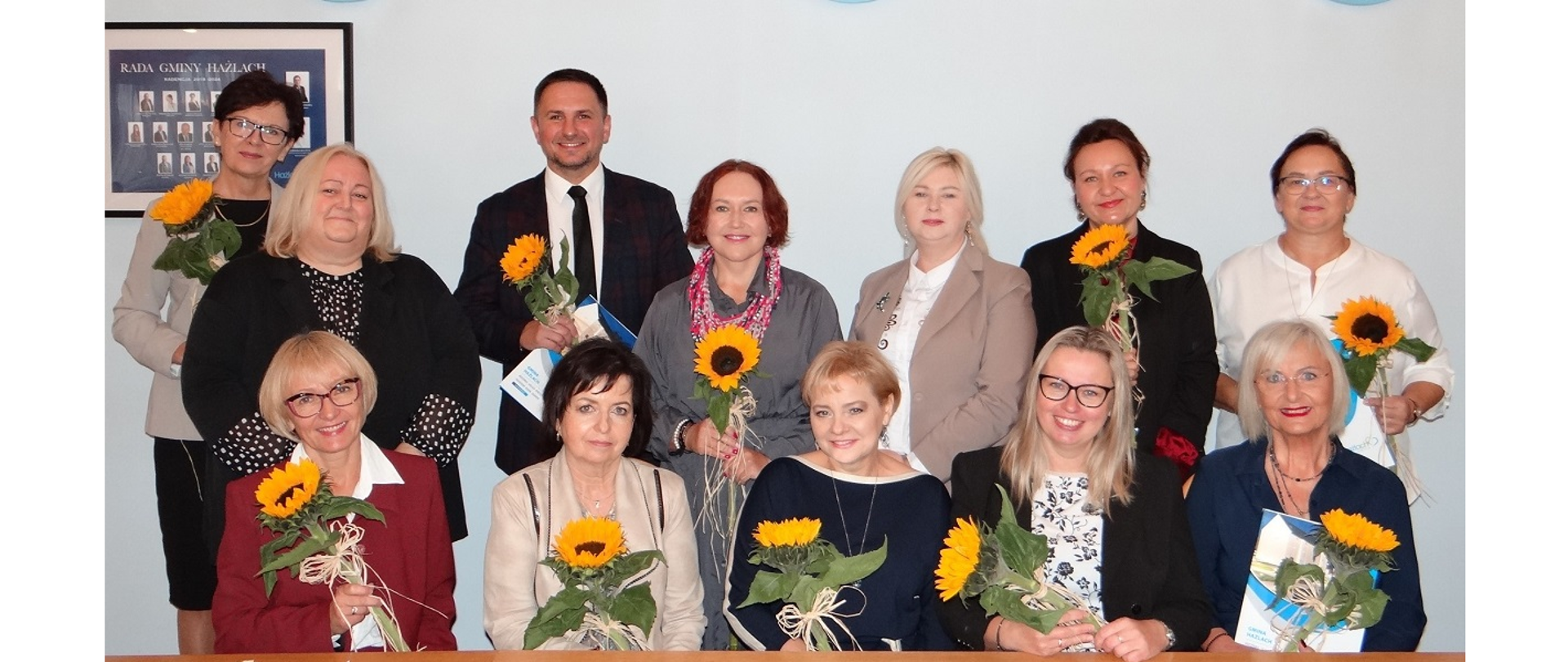 Wyróżnieni pedagodzy z przedstawicielami Gminy Hażlach. Osoby pozują wspólnie do pamiątkowego zdjęcia w sali Urzędu Gminy Hażlach. Nagrodzeni trzymają w ręku kwiaty